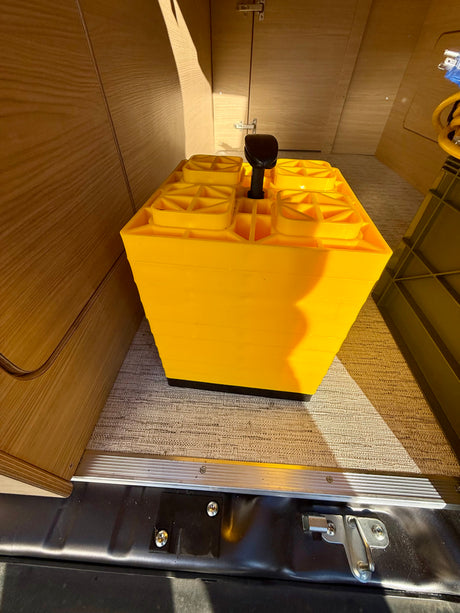 Bright yellow RV leveling blocks with a handle, stored inside an rv rental. Wood cabinets, carpet, and a floor latch are visible.