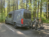 A gray RV rental van, a Mercedes Sprinter, with two mountain bikes on a rear rack, parked in a sunlit forest.
