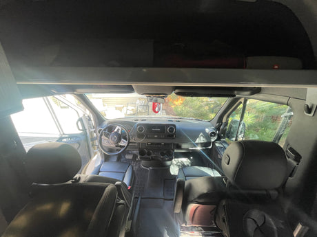 Interior view of an RV rental: front cabin with steering wheel, dashboard, and black leather seats, bright light from outside.