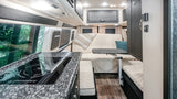 Modern RV rental interior: kitchen with stove, versatile living area, and large windows with green views.