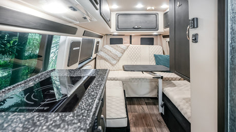 Modern RV rental interior: kitchen with stove, versatile living area, and large windows with green views.