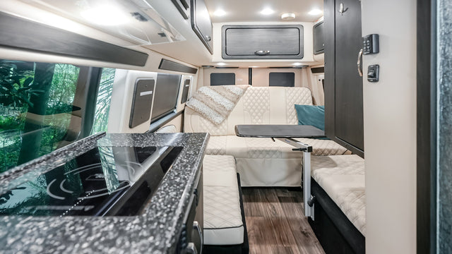 Modern RV rental interior: kitchen with stove, versatile living area, and large windows with green views.