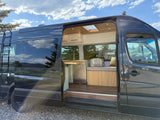 Explore in this custom dark grey camper van, an ideal RV rental. Side door open reveals a cozy, wood-finished interior.