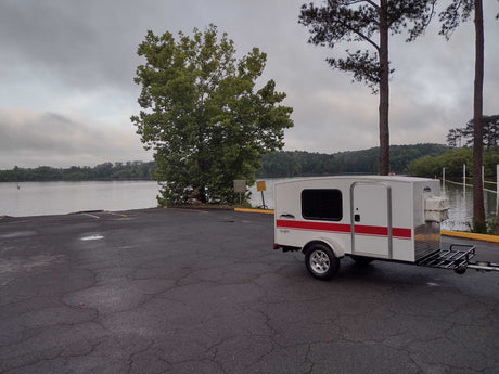 Compact white and red camper, perfect for an rv rental getaway, parked by a scenic lake and boat ramp on a cloudy day.