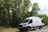 A white Ford Transit rv rental camper van, with its rear door ajar, parked by lush green trees in Utah on a sunny day.