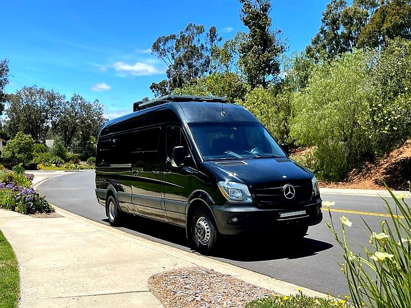 Black Mercedes-Benz Sprinter RV rental parked on a sunny road, surrounded by lush trees and flowers. Ideal for travel.