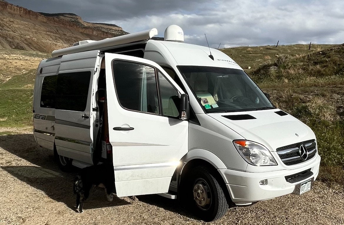 White Winnebago Era RV rental parked on a dirt road with its door open, a dog partially visible, against a mountain backdrop.