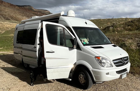 White Winnebago Era RV rental parked on a dirt road with its door open, a dog partially visible, against a mountain backdrop.