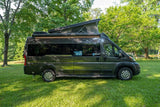 This dark green pop-top camper van, ideal for RV rental, is parked on a sunny grassy lawn amidst trees.