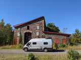 A grey RV rental camper van is parked on a road beside a unique stone building with a red roof and autumn foliage under a blue sky.