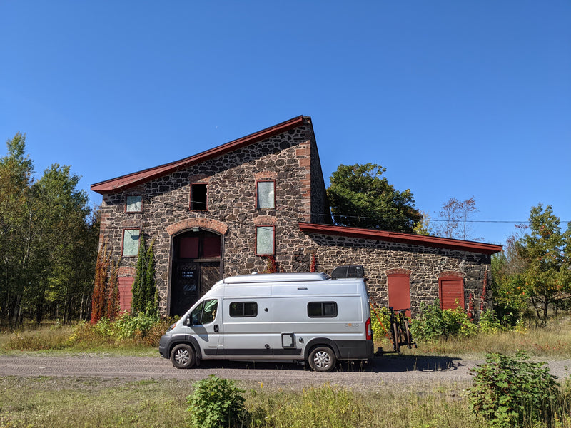 A grey RV rental camper van is parked on a road beside a unique stone building with a red roof and autumn foliage under a blue sky. — photo 4