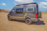 Adventure-ready gray Mercedes Sprinter RV rental van with off-road tires parked on a dirt lot near red rock formations.