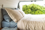Relax in your RV rental! A comfy bed with light linen bedding, pillows, and a lush green outdoor view from the window.