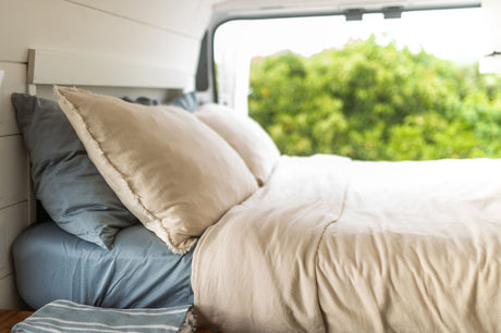 Relax in your RV rental! A comfy bed with light linen bedding, pillows, and a lush green outdoor view from the window.