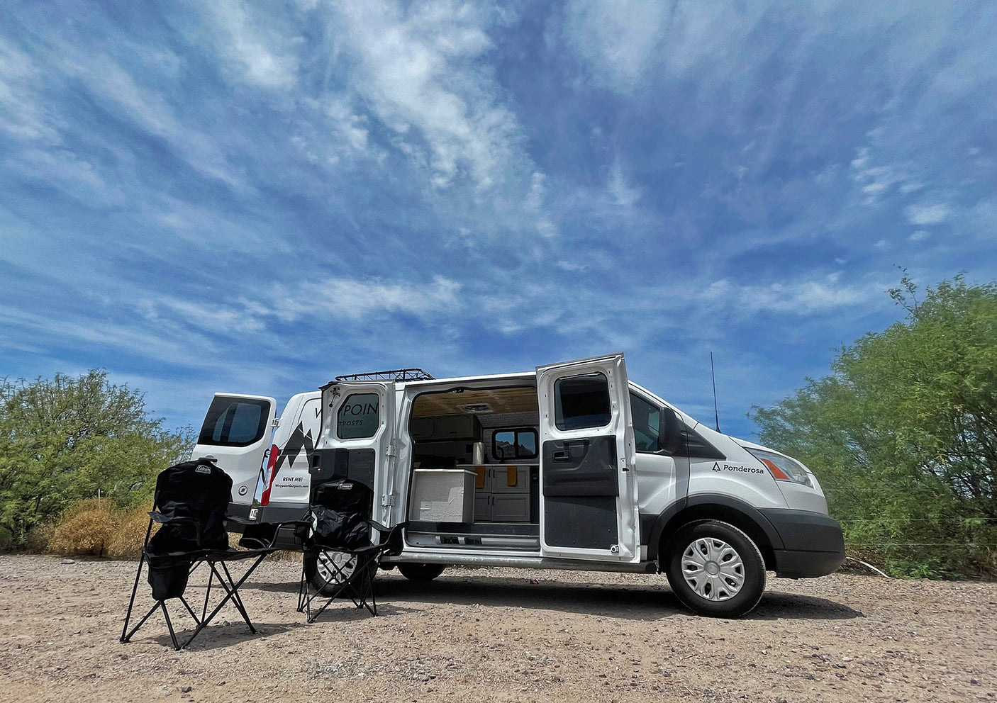 White RV rental van parked on a dirt road, side door open revealing a kitchenette, with two folding chairs set outside.