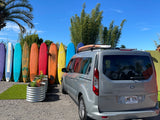 Gray Ford van, an RV rental, with surfboards on its roof, parked by a fence lined with colorful surfboards under a blue Hawaii sky.