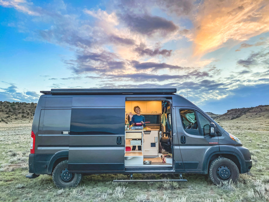 Your next adventure starts with an RV rental! A woman cooks inside her custom camper van, set in a vast, open landscape at dusk.