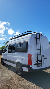 White Mercedes Sprinter RV rental van with roof rack & rear ladder, parked on gravel under blue sky.