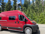 Bright red RV rental van parked roadside, amidst towering green trees, under a clear blue sky.