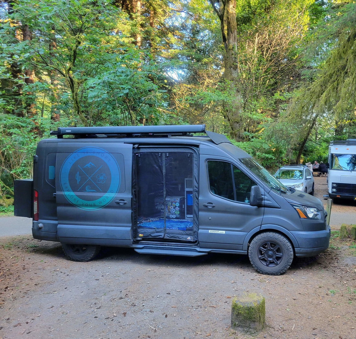 Dark gray RV rental camper van parked at a wooded campsite. Its side door is open