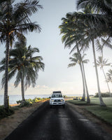 A white truck with roof tent on a coastal road lined with palm trees & ocean. Explore with an RV rental adventure!