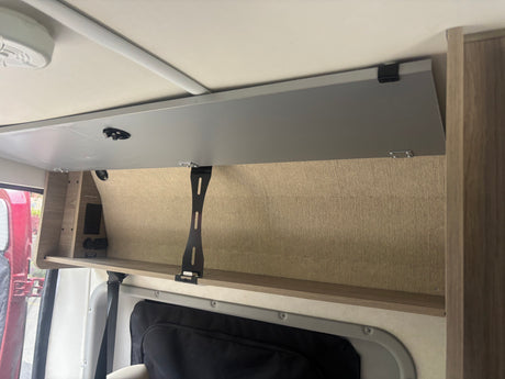 RV rental interior: Open overhead storage, a shelf with AC/USB/12V outlets, and a black support bracket in a camper van.