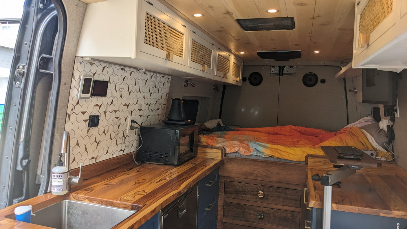 This custom RV rental interior features a kitchen with wooden countertops, tiled backsplash, and an adjacent bed for comfortable travel.