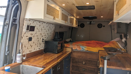 This custom RV rental interior features a kitchen with wooden countertops, tiled backsplash, and an adjacent bed for comfortable travel.