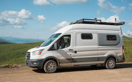 Scenic RV rental: a white camper van with a roof rack on a dirt road, mountains & sky in the background.
