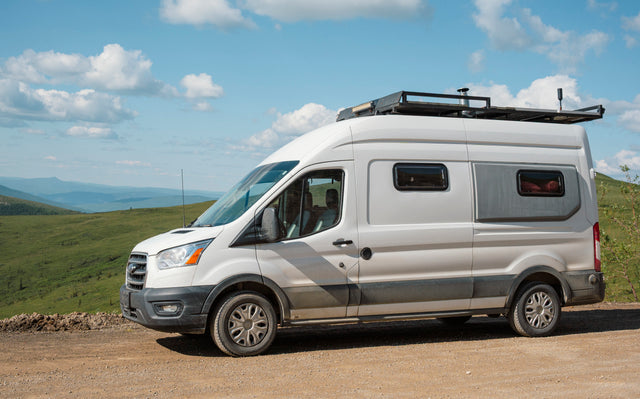 Scenic RV rental: a white camper van with a roof rack on a dirt road, mountains & sky in the background.