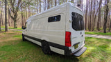 White Mercedes Sprinter RV rental parked on lush grass amidst tall trees. Perfect for your next nature adventure!