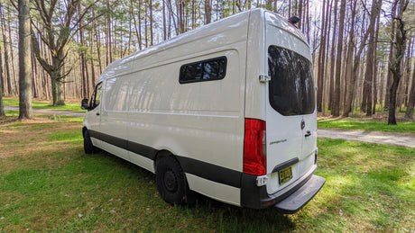 White Mercedes Sprinter RV rental parked on lush grass amidst tall trees. Perfect for your next nature adventure!