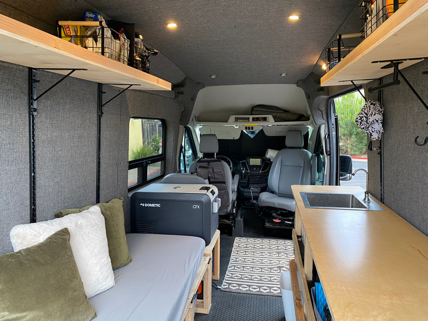 Cozy RV rental camper van interior with bed, Dometic fridge, kitchen sink, wooden shelving, and front seats visible.