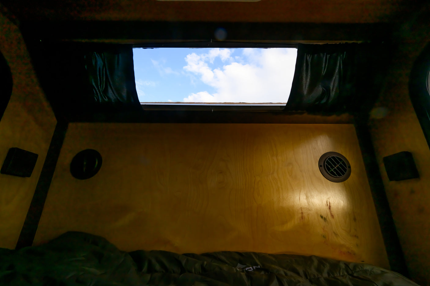 Inside an RV rental: a cozy wooden interior with a window revealing a blue sky and clouds, a bed, and wall vents.