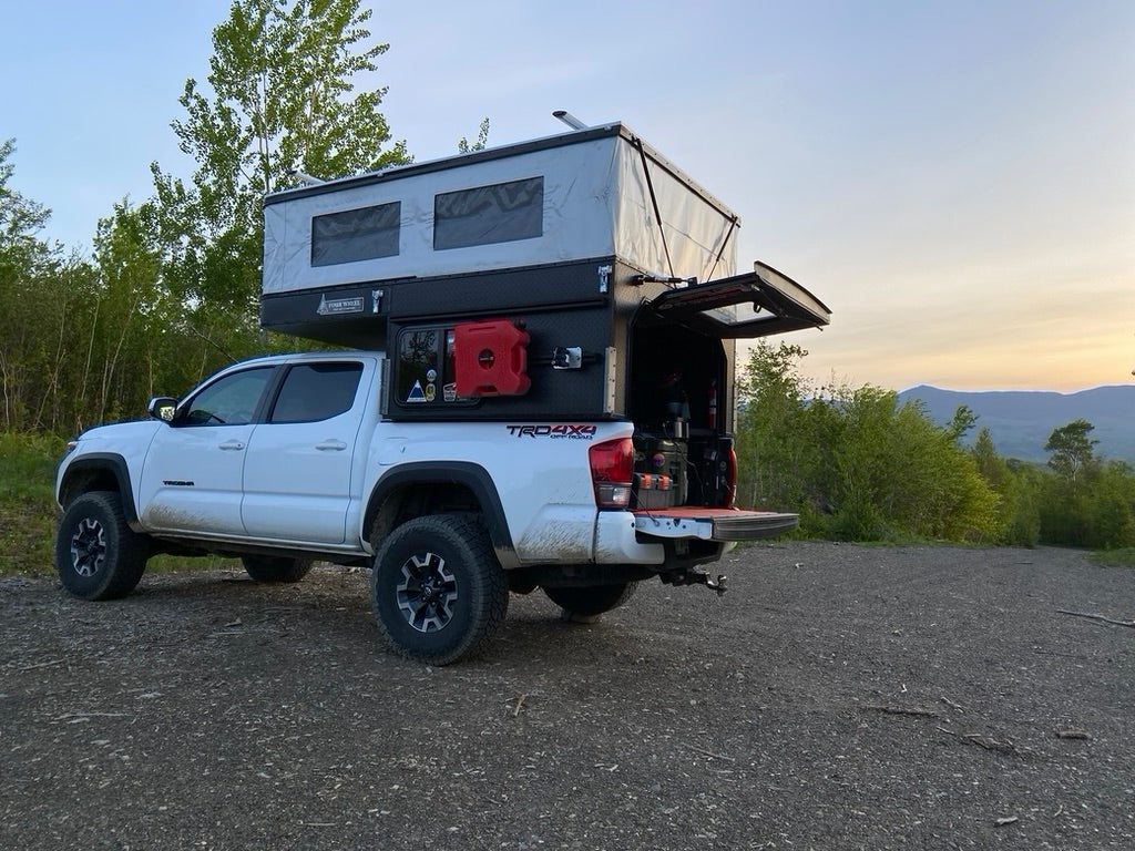 White Toyota Tacoma with pop-up truck camper parked on gravel. Ideal RV rental for scenic outdoor adventures.