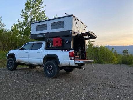 White Toyota Tacoma with pop-up truck camper parked on gravel. Ideal RV rental for scenic outdoor adventures.