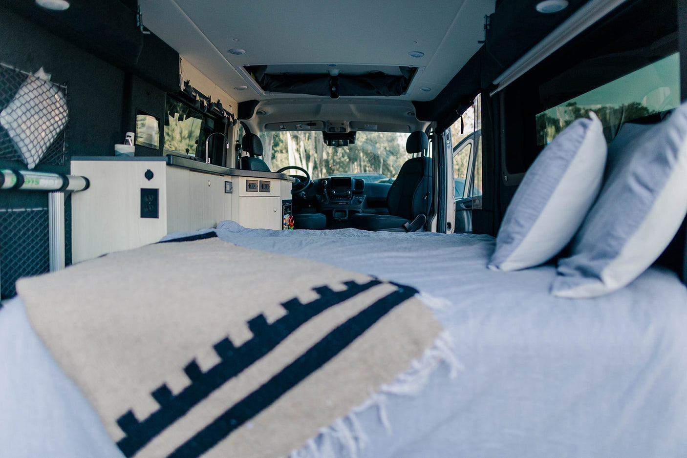 Spacious RV rental interior: comfortable bed with blanket, compact kitchen, and front driver's cabin visible.