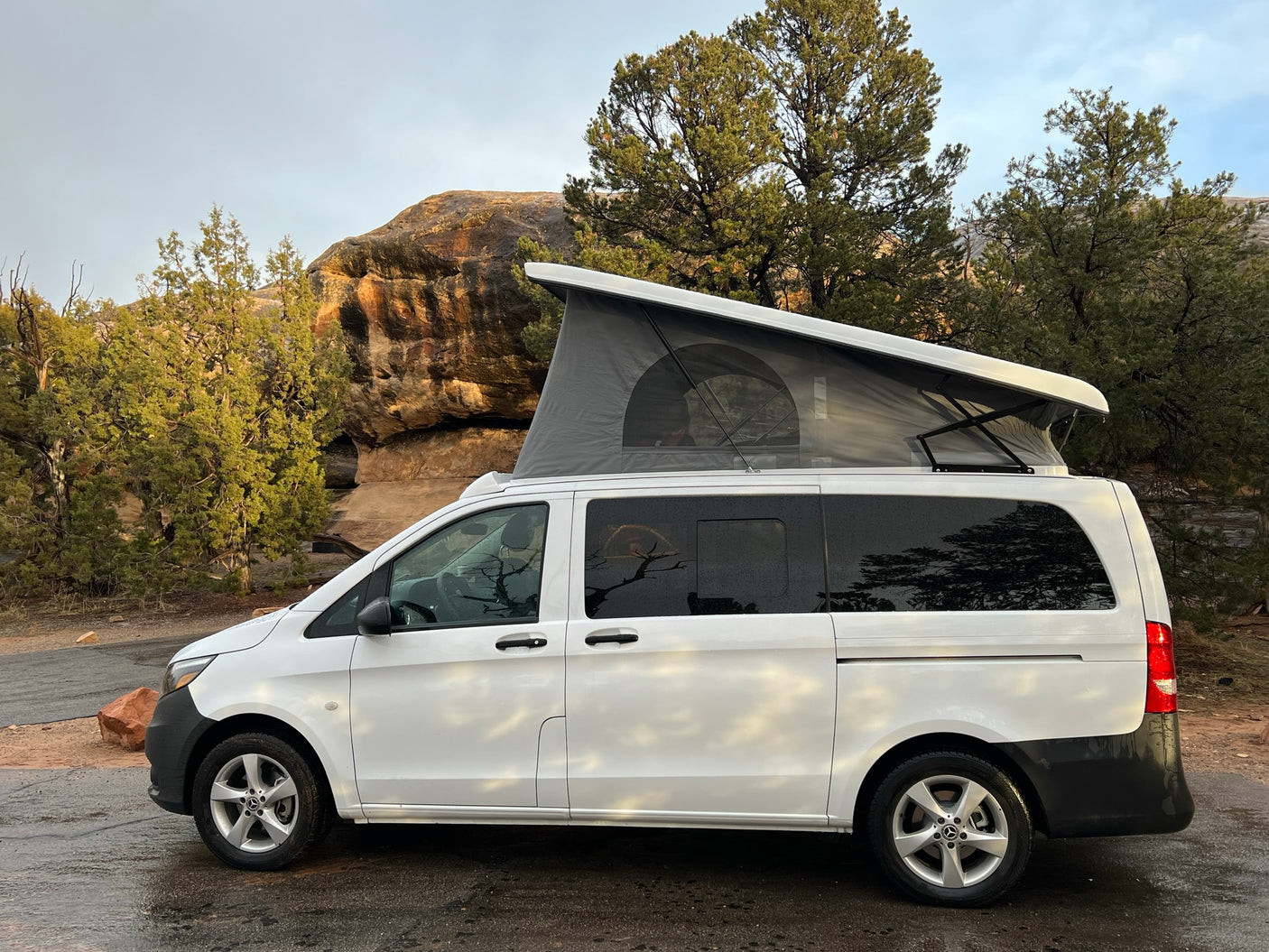 White Mercedes camper van with pop-top open, parked in a rocky, forested area. This RV rental is ready for your next adventure.