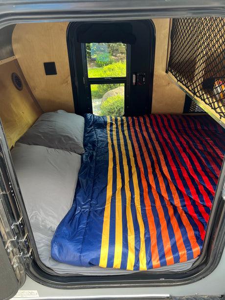 RV rental camper interior: cozy bed with vibrant striped comforter, pillows, and a refreshing window view of nature.