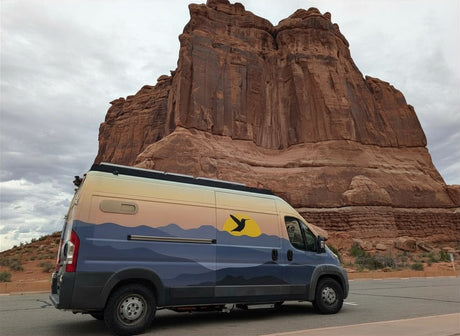 This scenic rv rental shows a custom camper van by towering red rock formations under a cloudy sky. Great for exploring national parks.