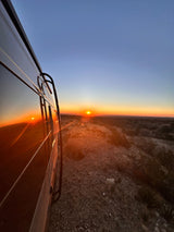 An RV rental reflects a vibrant desert sunset, parked on a rocky hill overlooking a vast landscape.