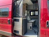 Open side door reveals a red RV rental's interior: compact fridge, bed/seating, and storage in a modern camper van.