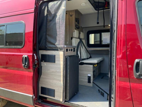 Open side door reveals a red RV rental's interior: compact fridge, bed/seating, and storage in a modern camper van.