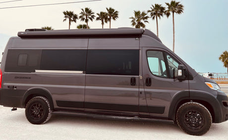 Gray RV rental camper van, "SEQUENCE" model, parked on a white shore with palm trees & ocean views. Perfect for coastal trips.