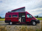 This red pop-top RV rental is parked on a gravel shoulder, its open side door revealing the interior against fields and sky.
