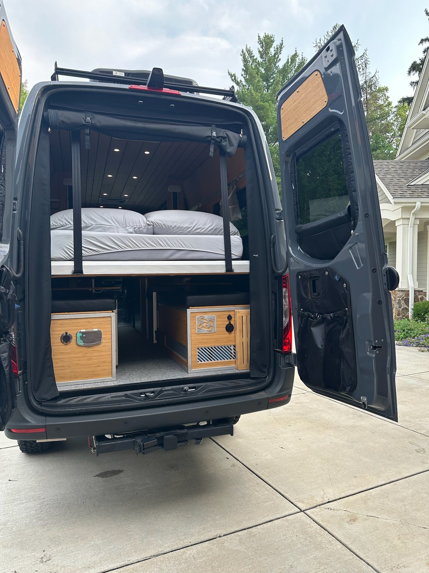 Rear view of an RV rental camper van with open doors, showcasing a raised bed, wood ceiling, and custom storage.