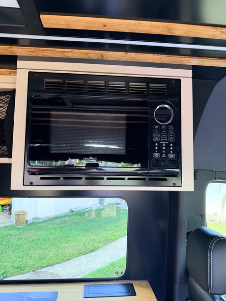 Inside an RV rental: a black HIGH POINTE microwave oven integrated into custom light cabinetry above a window, ideal for travel.