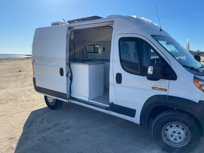  parked on a paved lot amidst a pine forest and rocky hill.|White RV rental camper van parked by a serene lake under a clear blue sky. Perfect nature escape!|White Ram ProMaster camper van RV rental on a sandy beach. Inside shows a modern kitchen. Perfect for van life travel.|Ready for an **RV rental** adventure! White camper van with open back revealing a cozy interior — photo 4