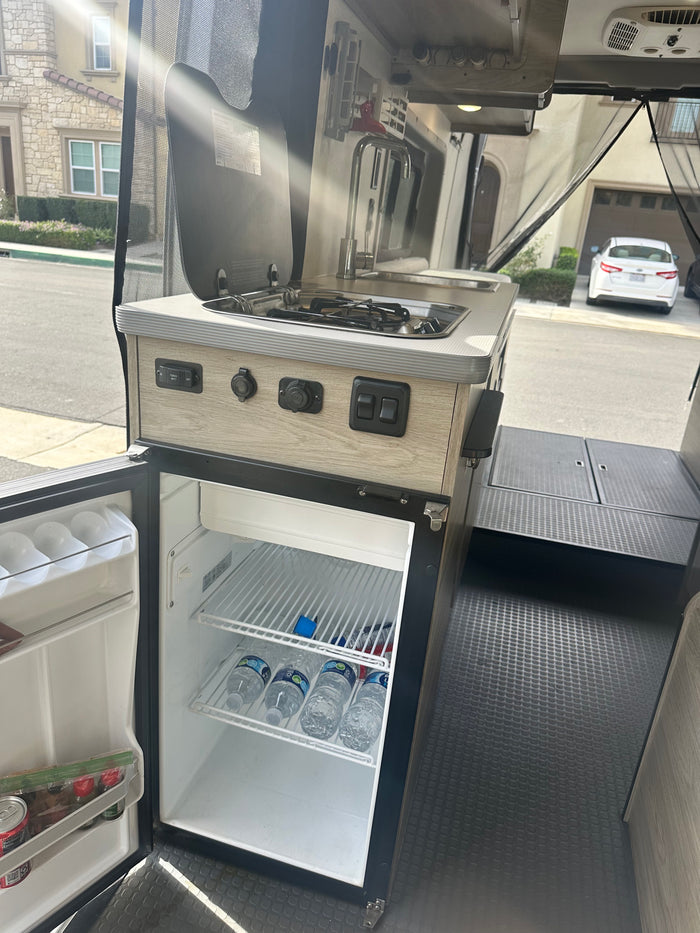 This RV rental's compact kitchen boasts a sink, stove, and a stocked fridge, ready for any adventure.