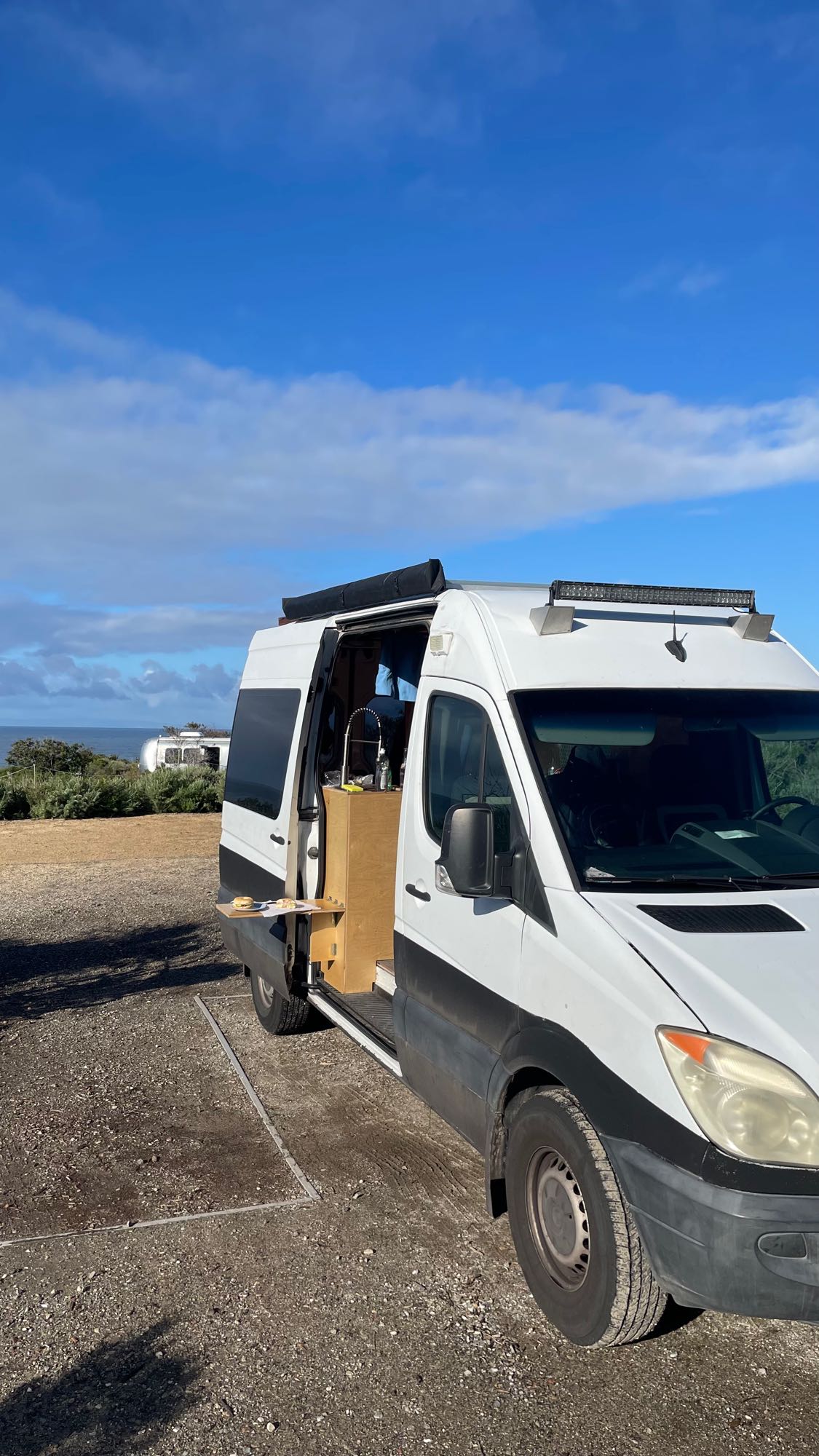 Scenic RV rental: White camper van with open kitchen, parked on gravel with ocean views. Perfect mobile setup.